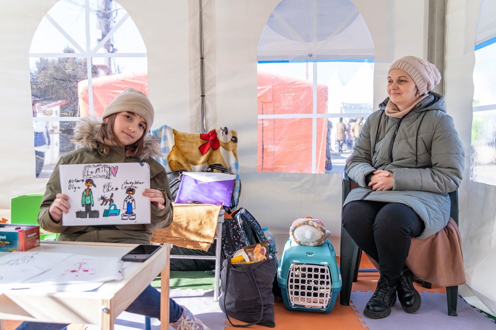 A nine-year-old Ukrainian girl holds a drawing of her family as she sits in a learning hub with her mother and cat (in blue basket) in Romania. A nine-year-old Ukrainian girl holds a drawing of her family as she sits in a learning hub with her mother and cat (in blue basket) in Romania.