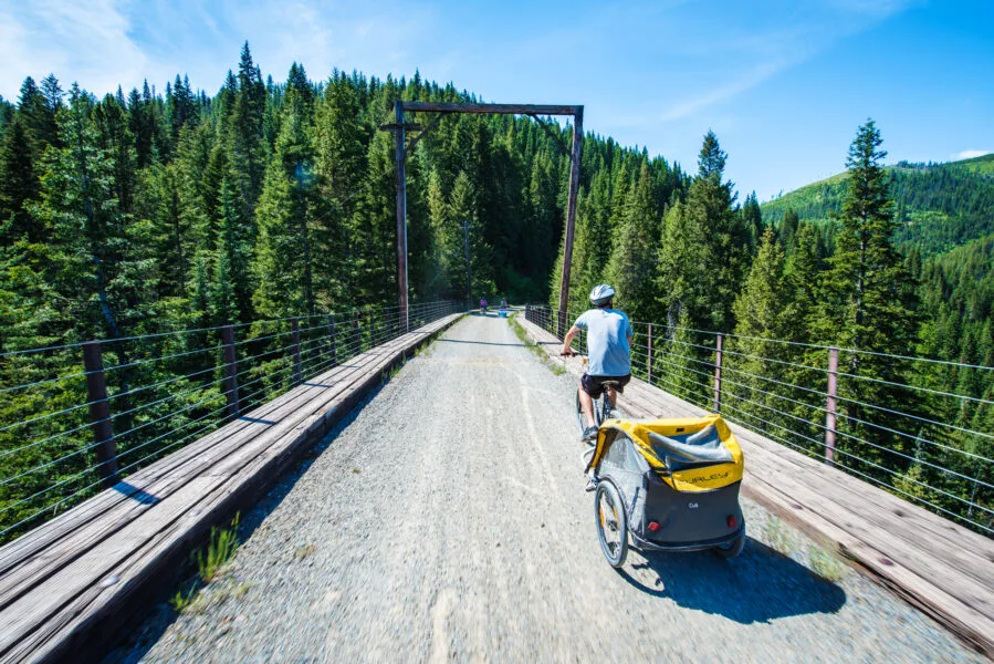 Man biking while towing a yellow children bike trailer along one of the trestle bridges on the historic Route of the Hiawatha, a gravel rail-trail in North Idaho.