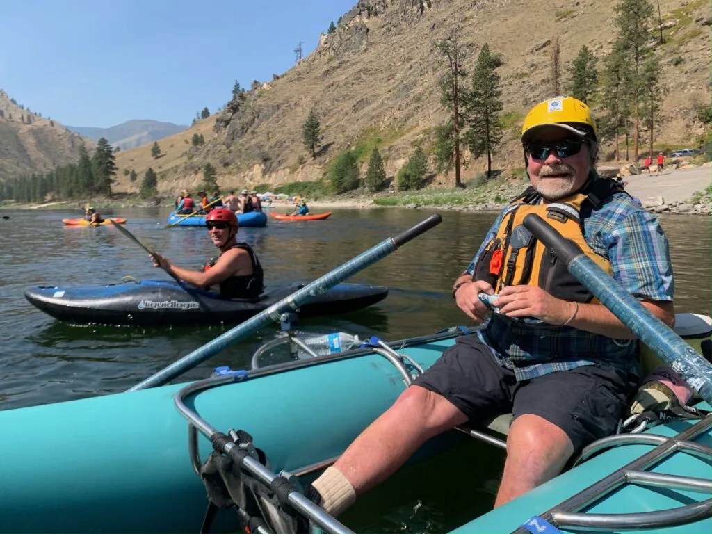 Man wearing a PFD and safety helmet sitting in a teal-blue inflatable raft, resting his paddles, on the Salmon River.