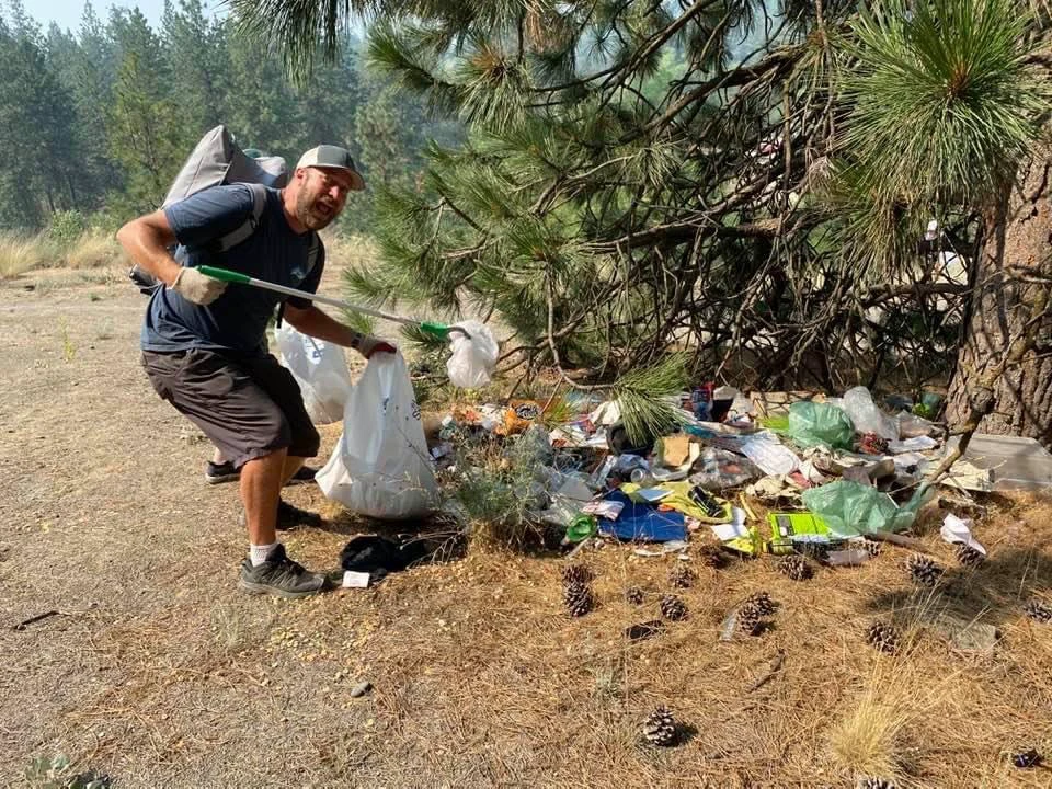 Man using a gripping tool to pick up trash along the banks of the Spokane River and put garbage into trash bag.