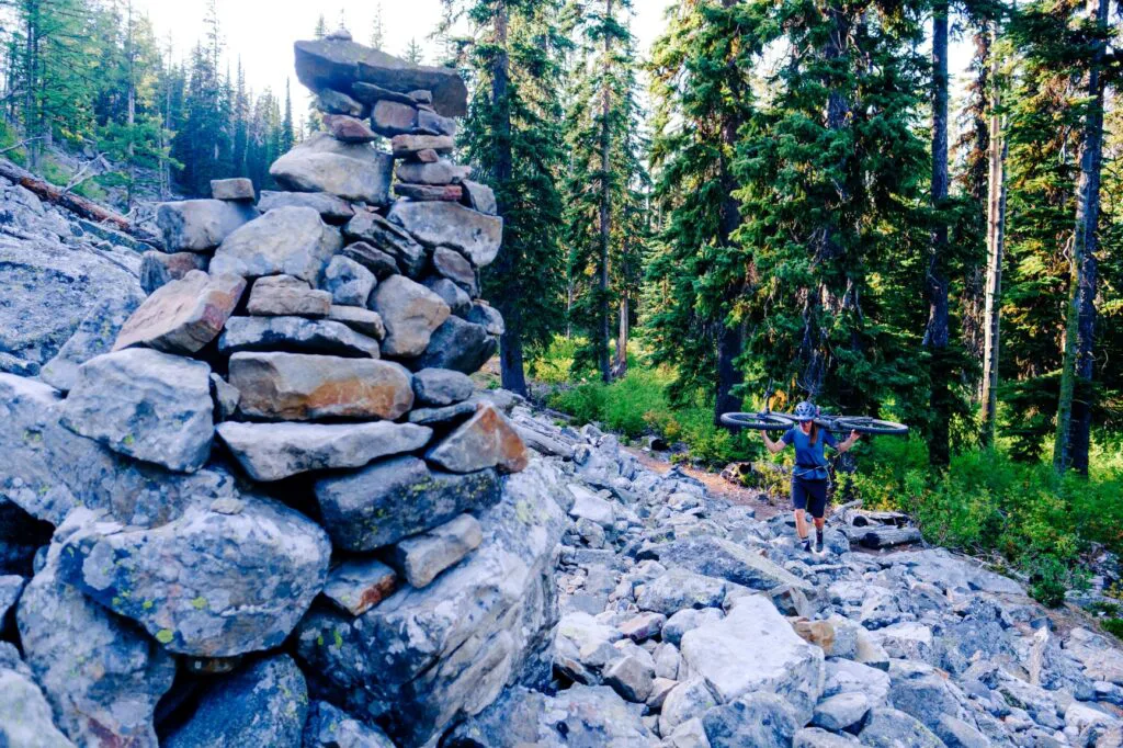 Woman carrying her mountain bike over her shoulders as she hikes over a rocky trail across a talus slope.