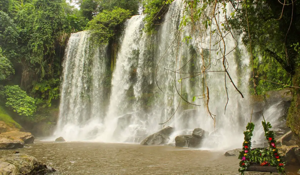 A tall waterfall with a swing in front of it in the middle of a lush jungle in Phnom Kulen, Cambodia