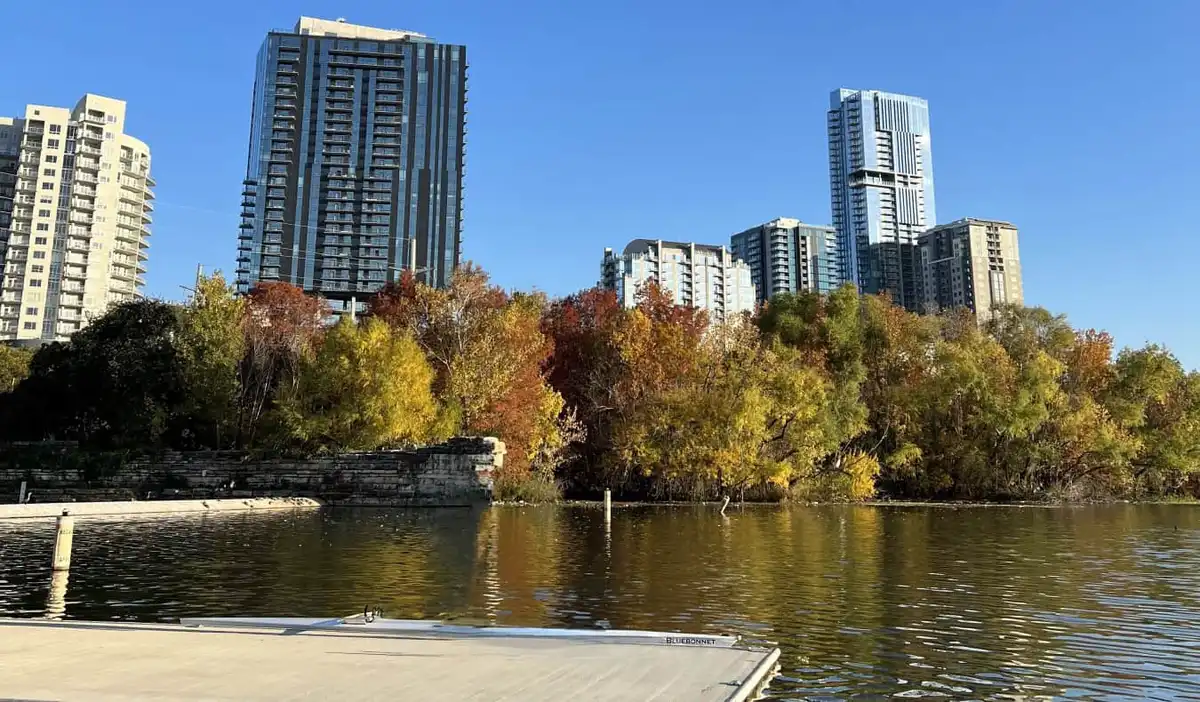 A lake in Austin, Texas, with a a dock in the foreground, trees filled with orange and yellow leaves on the edge of the lake, and a skyline of skyscrapers in the background