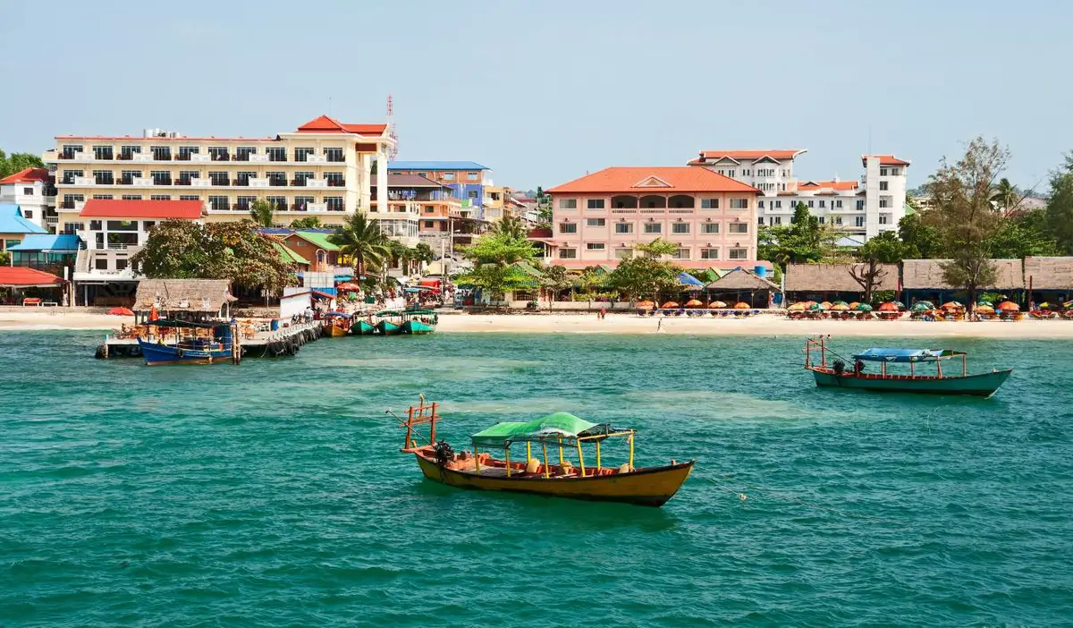 A city beach in Sihanoukville, Cambodia, with colorful hotel buildings, loungers on the beach, and boats docked in the water